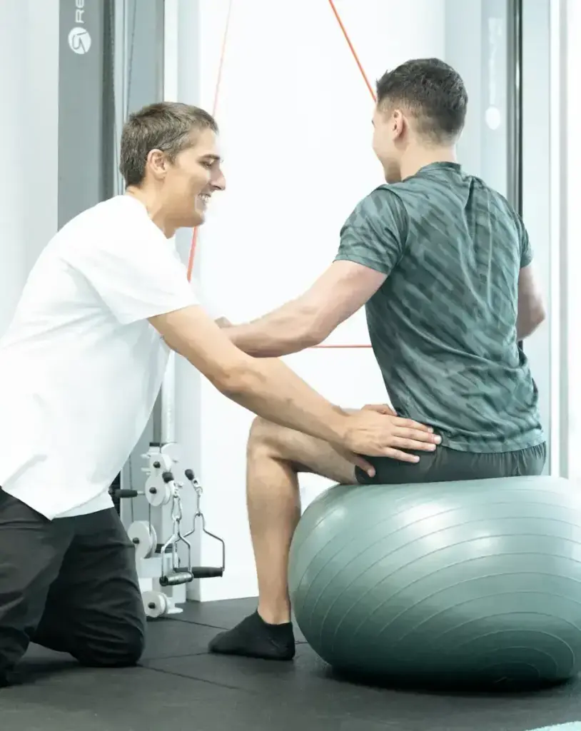 Physiotherapeut unterstützt Patienten bei klassischer Krankengymnastik mit Übungen auf dem Gymnastikball im Trainingsraum