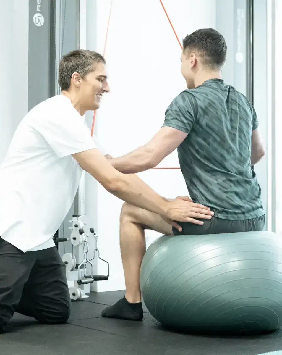 Physiotherapeut unterstützt Patienten bei klassischer Krankengymnastik mit Übungen auf dem Gymnastikball im Trainingsraum
