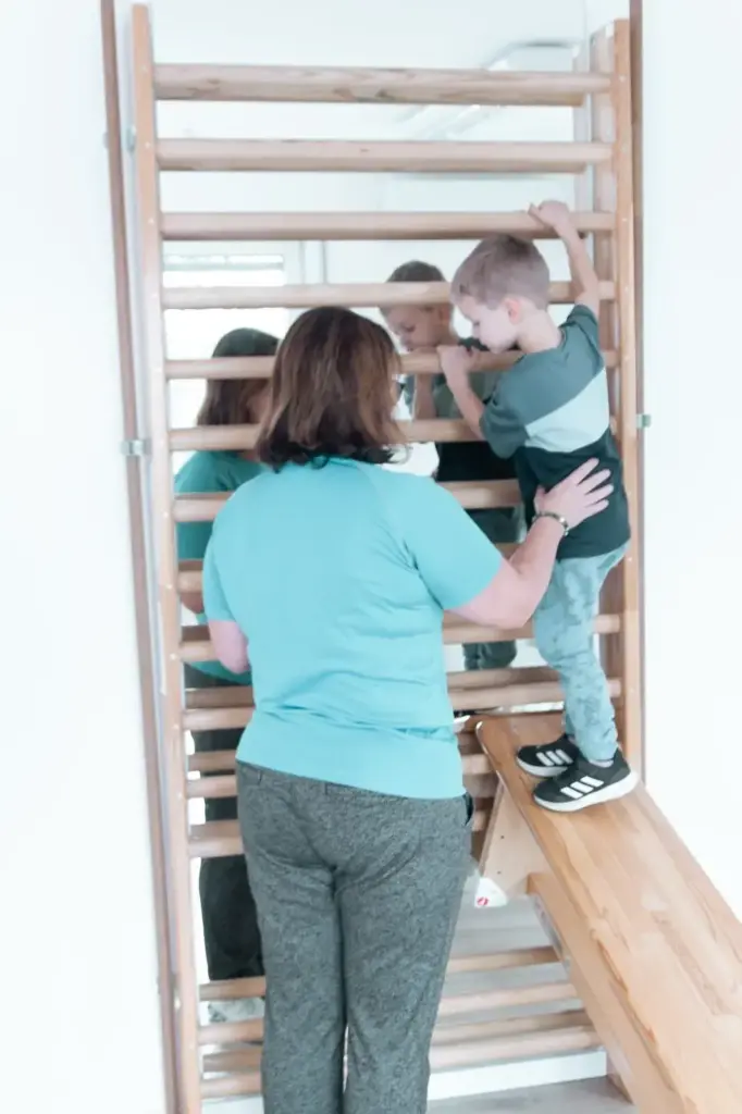 Therapeutin unterstützt ein Kind beim Klettern an der Sprossenwand in der Kinderphysiotherapie zur Förderung von Kraft und Koordination